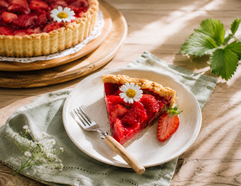 A slice of glossy strawberry tart with agar glaze on a pale ceramic plate, with the remaining tart and white daisy-like flowers in the background.